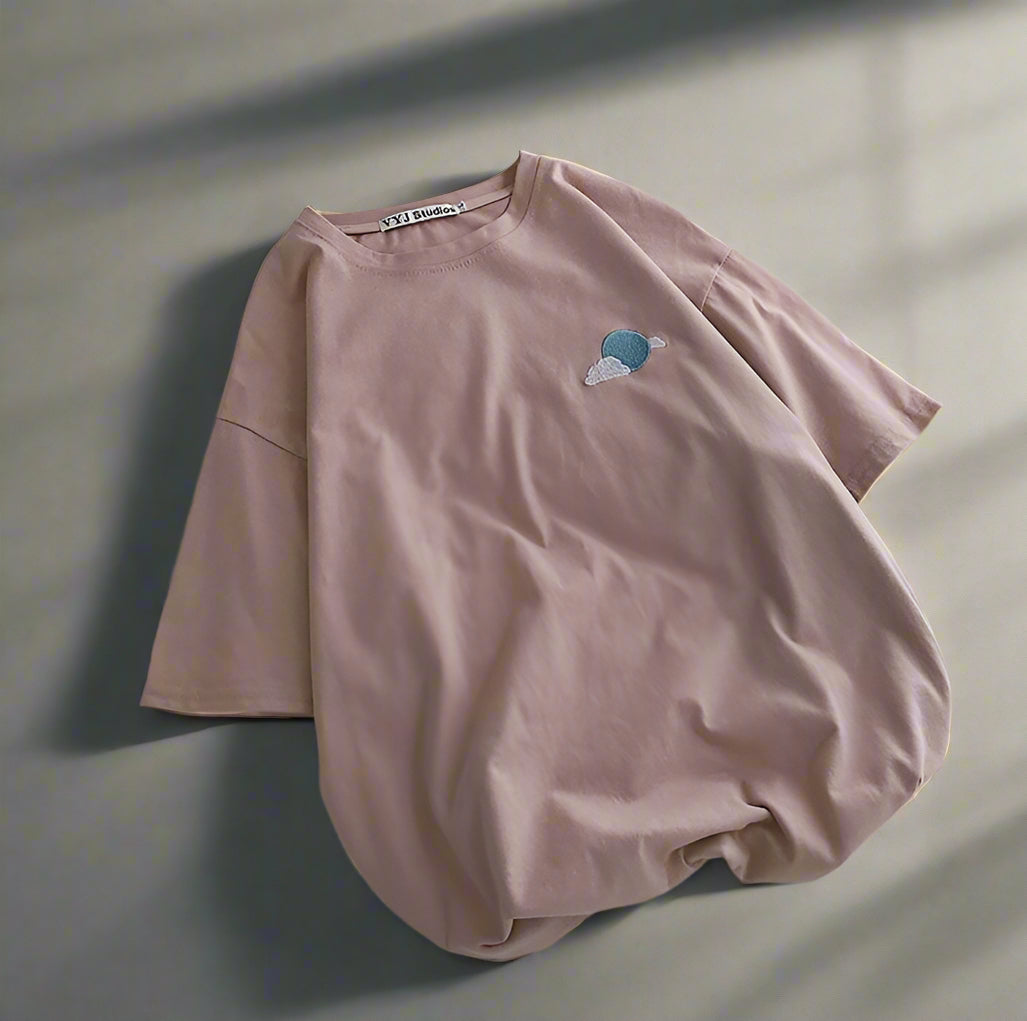 Pink t-shirt with a small cloud design on a white surface with shoes and a magazine.