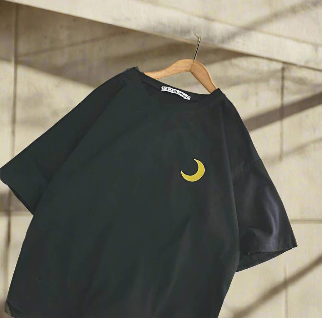 Black t-shirt with a yellow moon design on a white surface with shoes in the background.